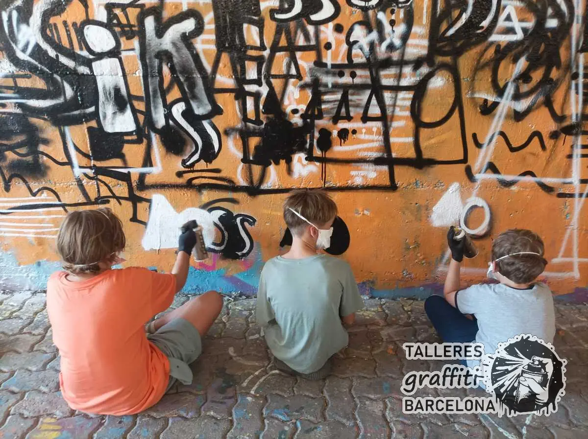 Participantes con mascarilla pintando letras negras sobre un muro naranja en un curso de graffiti en Barcelona